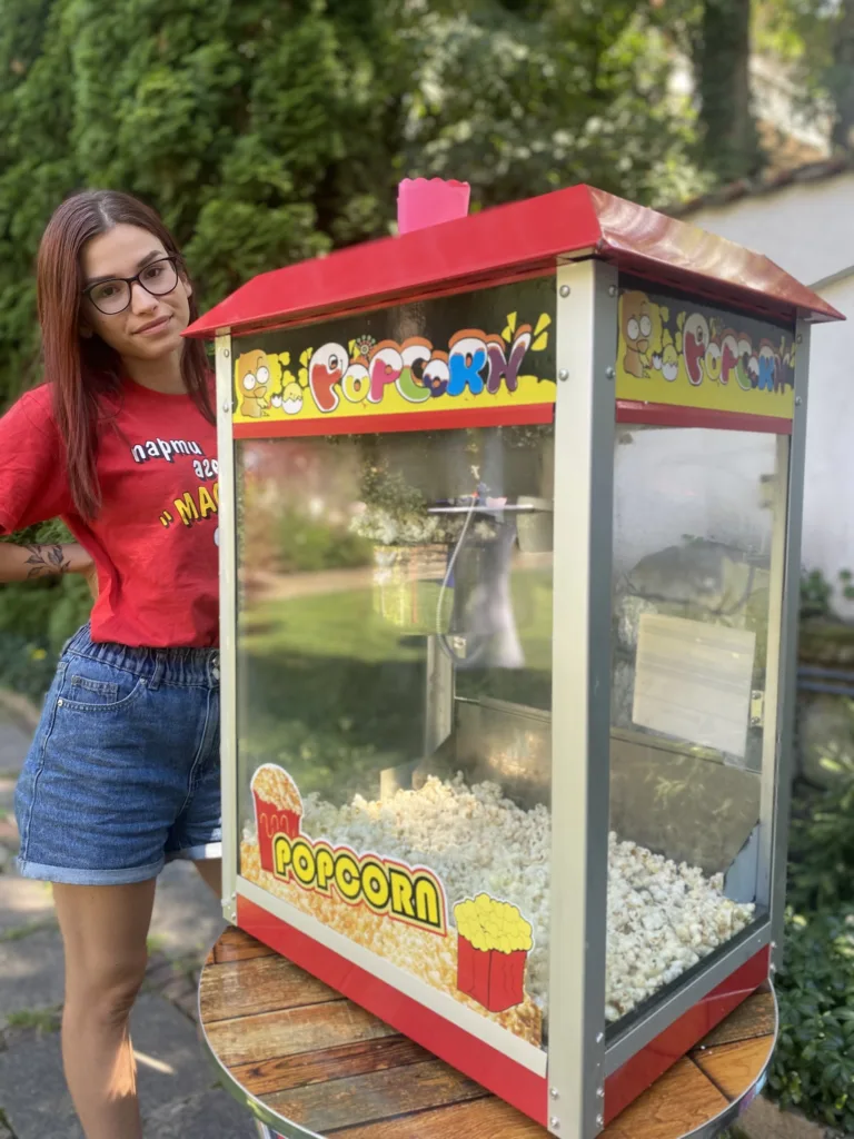 Аниматорка с очила и червена тениска стои до машина за пуканки с надпис Popcorn.