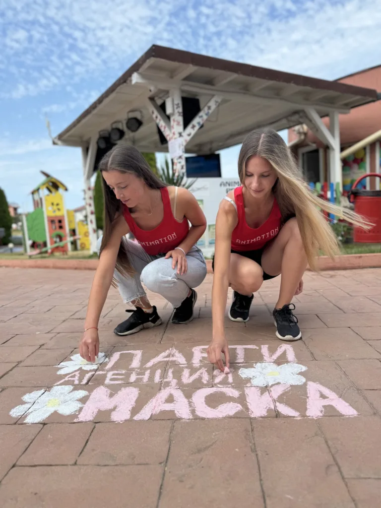 Две аниматорки рисуват с тебешир по плочките надпис Парти агенция Маска, зад тях навес и площад.