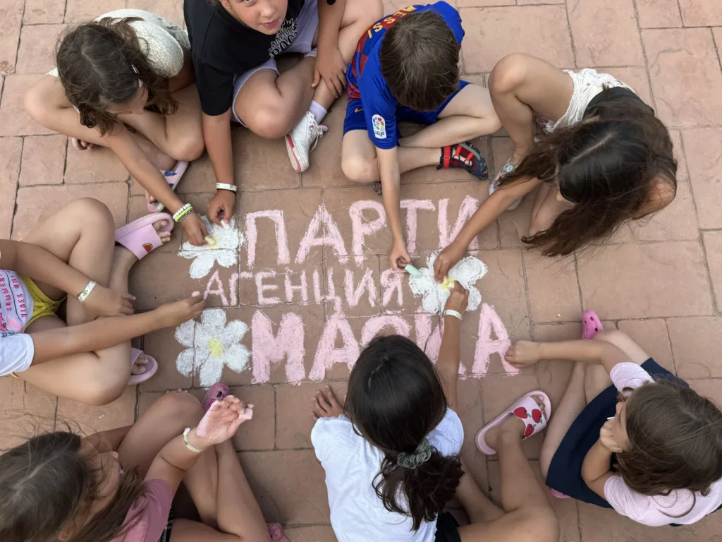Деца с бели цветя в ръце седят около тебеширен надпис Парти агенция Маска, гледано отгоре.
